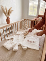 Pochon de naissance J1 et tenue de bébé en gaze de coton disposés sur un lit bébé, ambiance terracotta et bois.