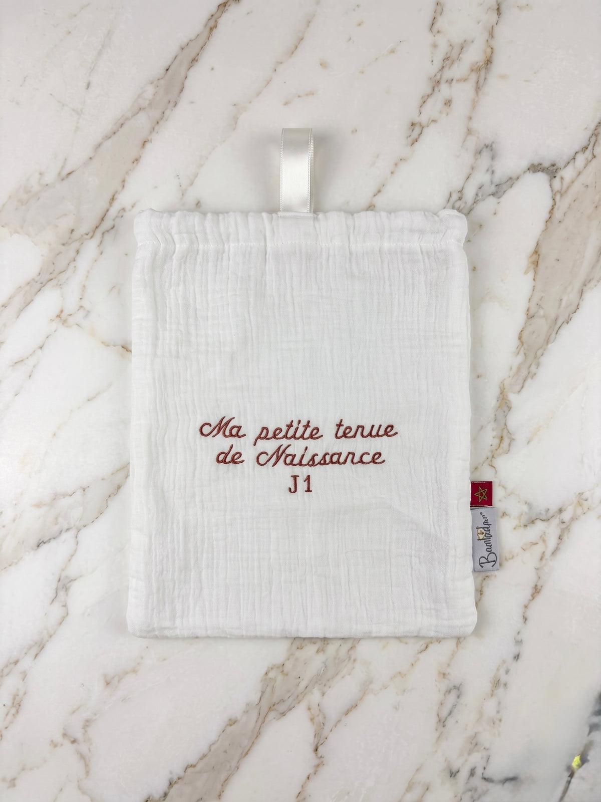 Pochon de naissance blanc brodé terracotta J1 posé sur un plan de travail en marbre blanc et doré.